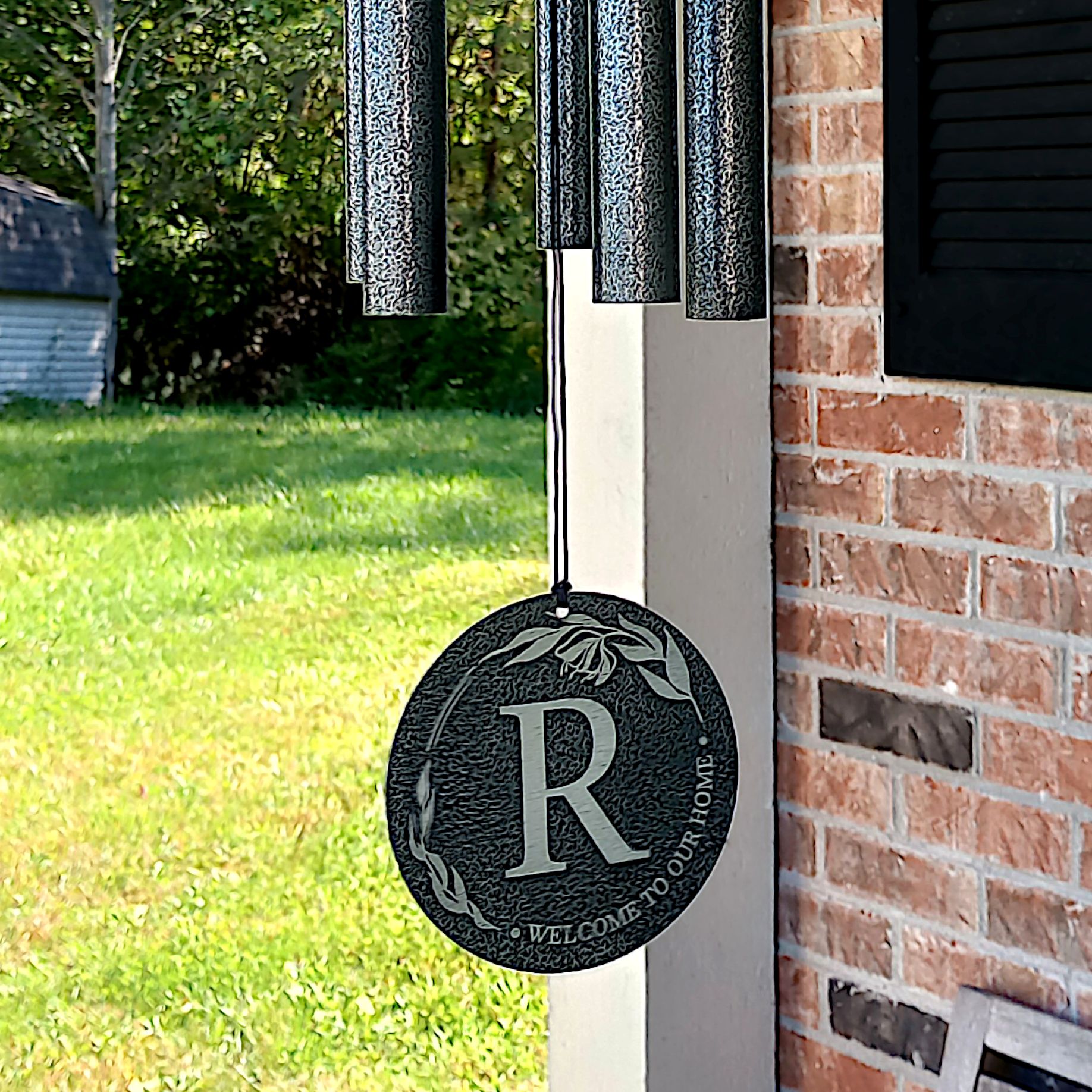 A black, powder-coated personalized wind chime hanging over the porch of a red brick house with black shutters. The sail features the letter R surrounded by a leafy pattern and the words "Welcome to our home".