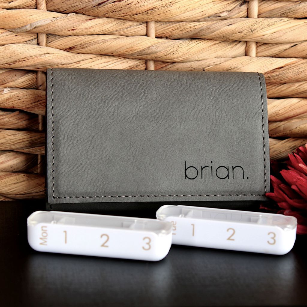 A grey leatherette pill case personalized with the name Brian. Two of the seven daily pill organizers are displayed in the foreground on a black countertop against a wicker wall.
