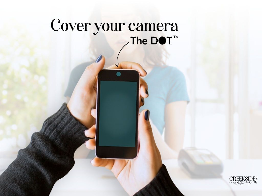 A pair of hands holding a mobile phone while standing at a retail checkout counter. The phone's camera is covered by The DOT™, which is blocking the camera's view of everything but the color of The DOT™ that is applied (in this case, teal).